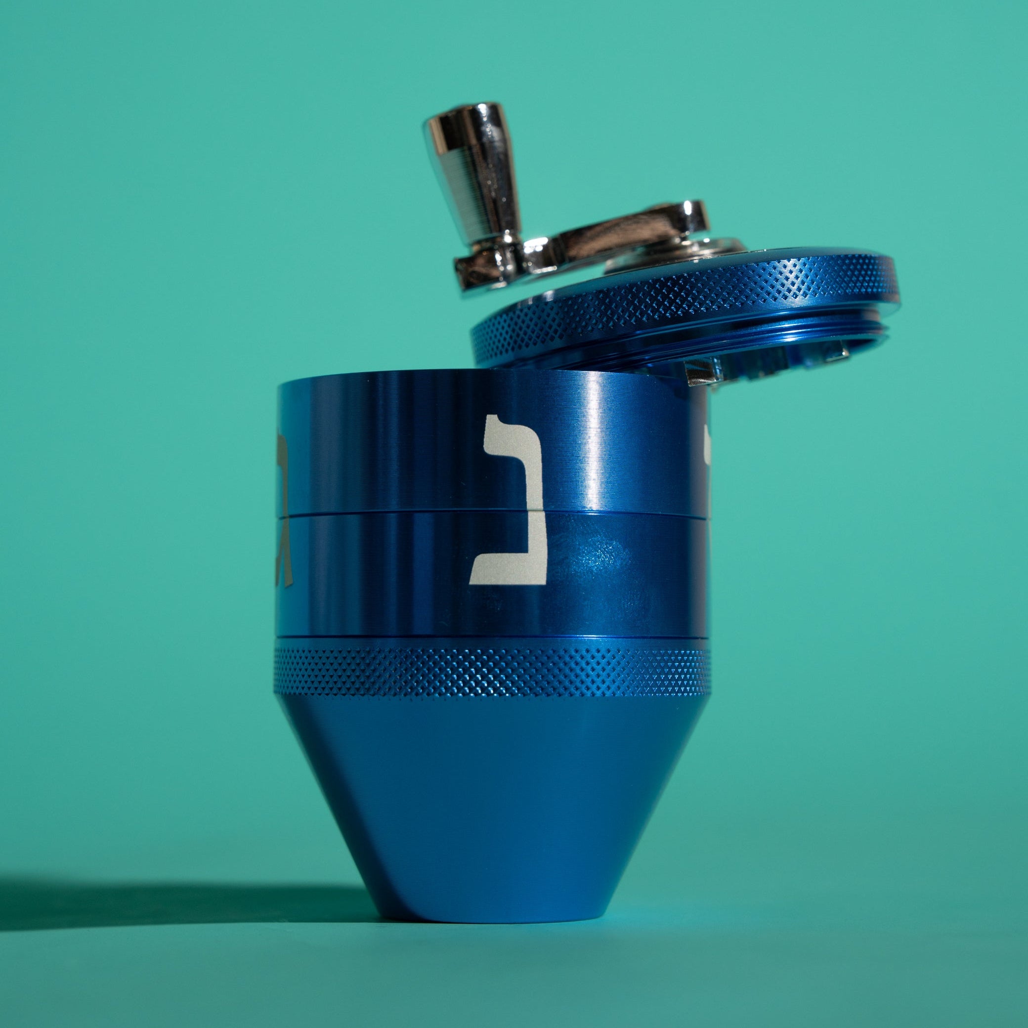 A metallic blue manual herb grinder with a silver metal handle, shown on a solid teal background.
