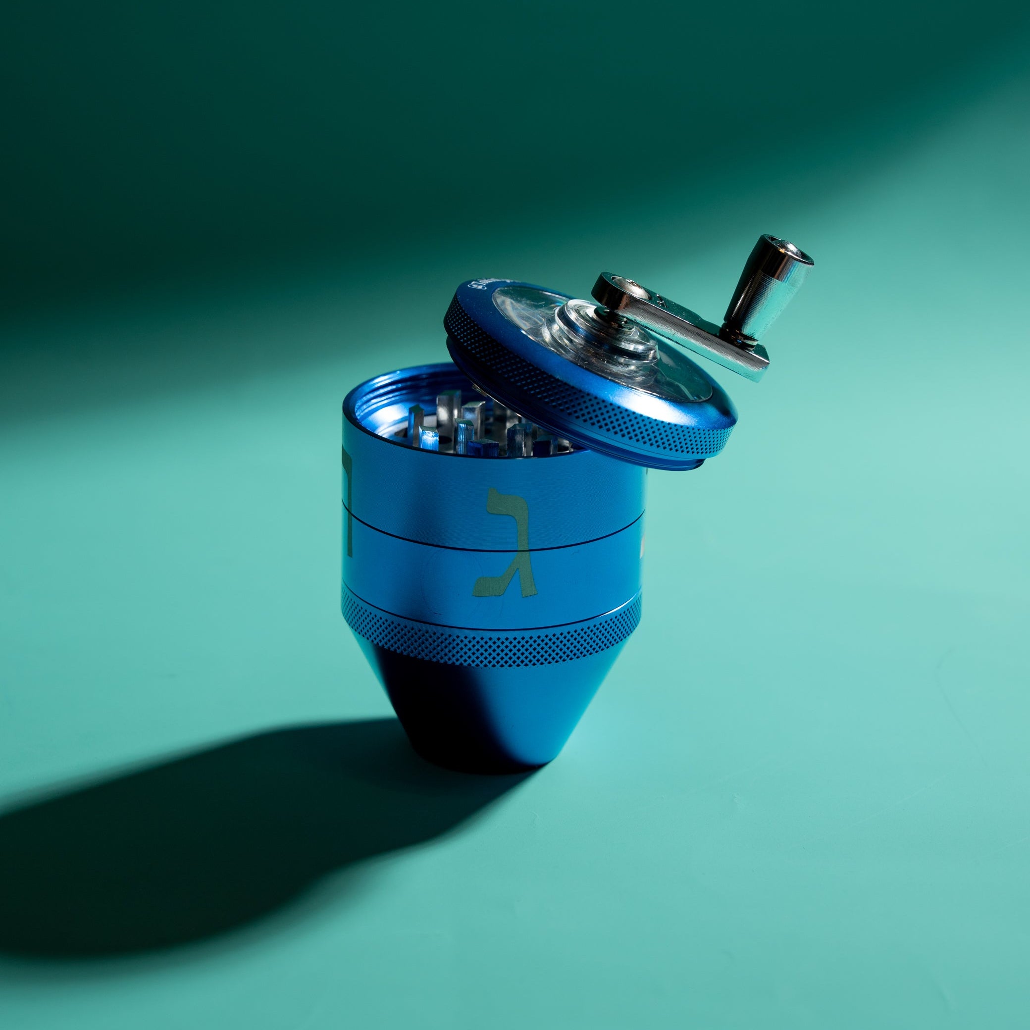 A metallic blue manual herb grinder with a silver handle, shown open on a teal shadowed background.