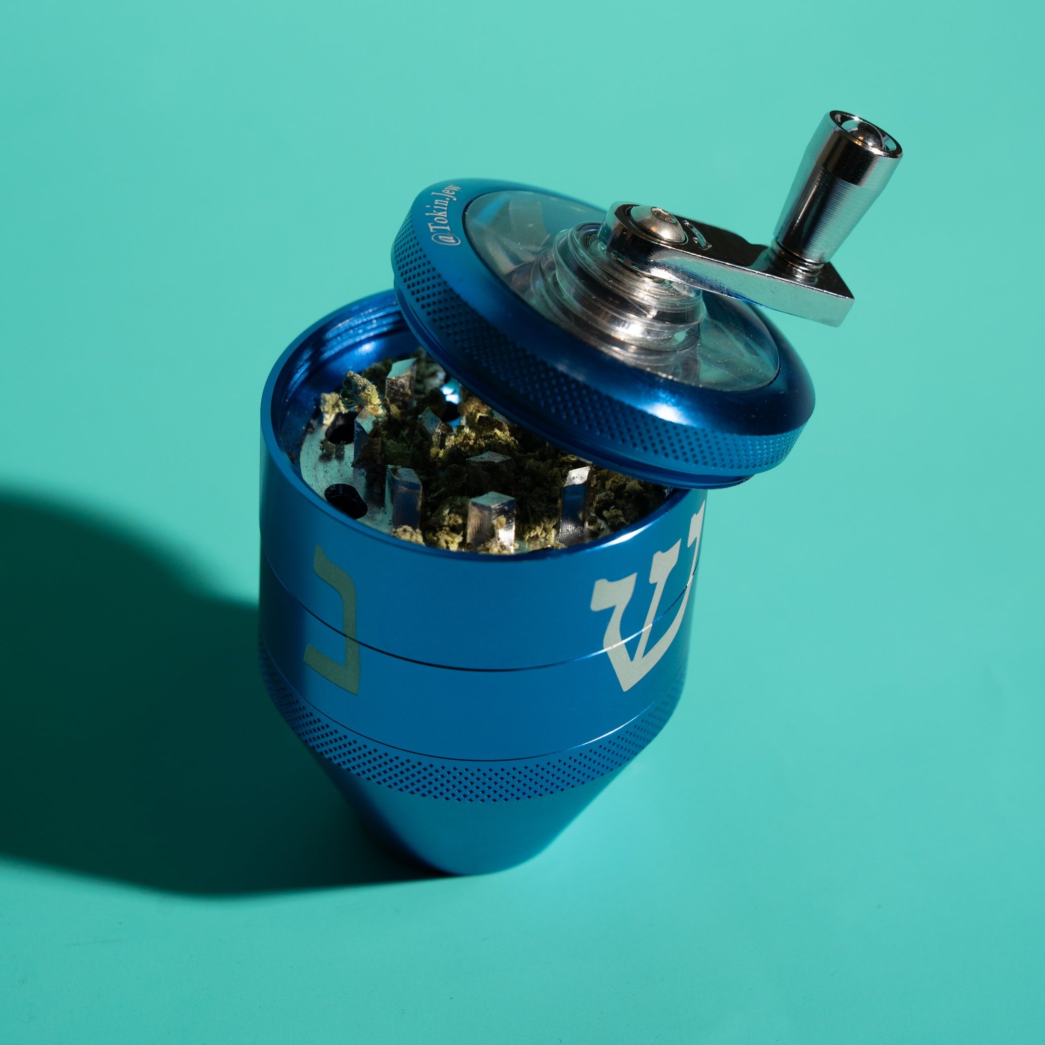 A blue metal herb grinder with a silver handle and lid, shown open and filled with dried herbs on a solid teal background.