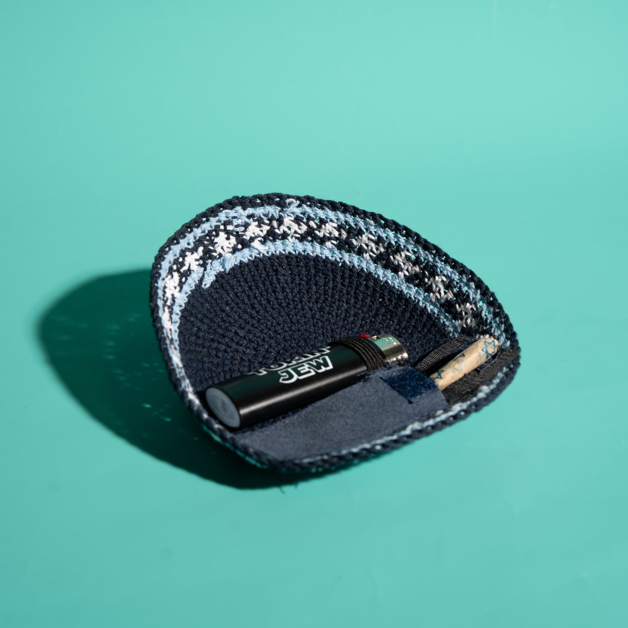 A knit kippah in navy blue with a white and light blue patterned trim, shown on a solid teal background. Inside the pouch, a black lighter and a small Tokin' Jay are placed.