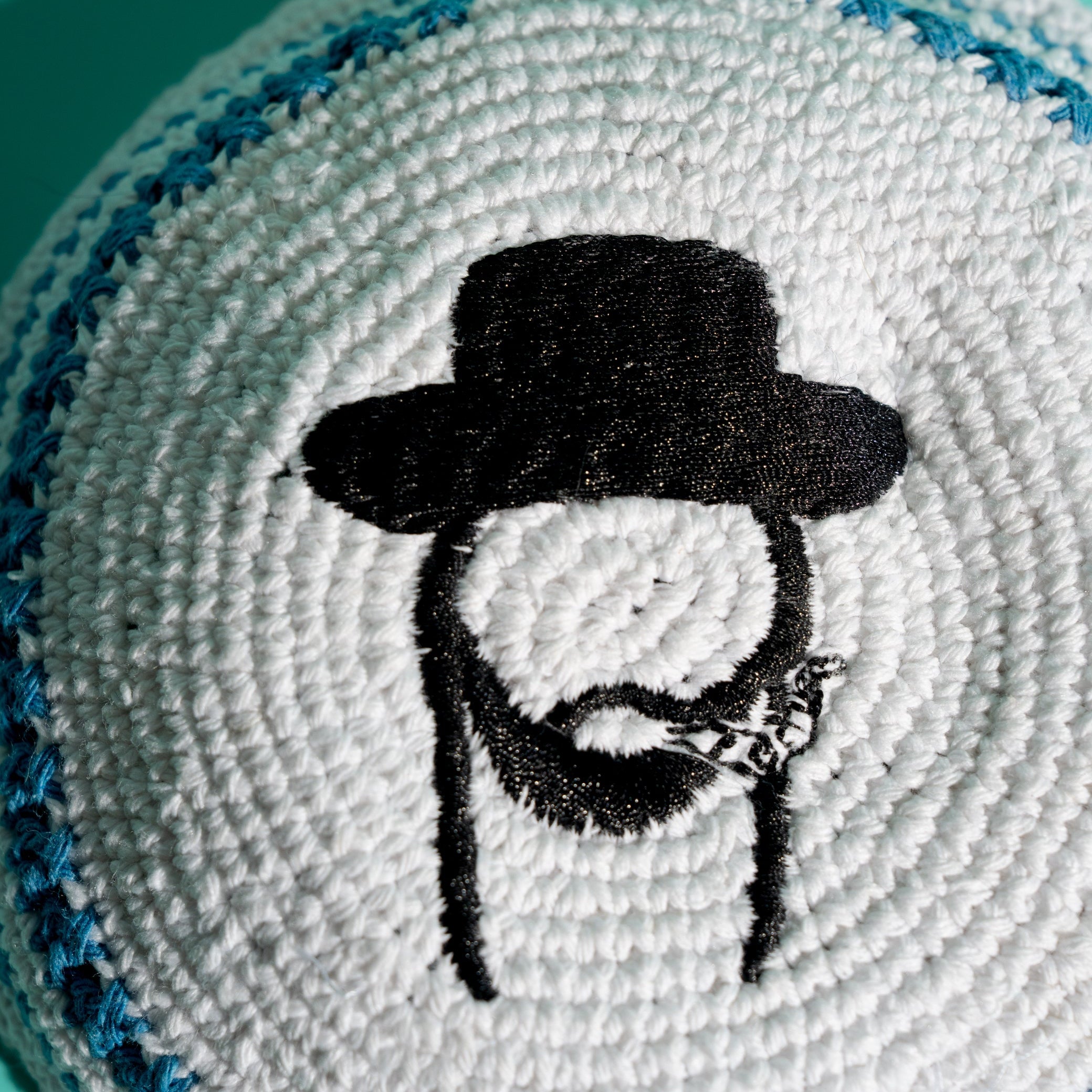 A close-up of a white knit kippah, featuring a black embroidered portrait of a Rabbi smoking a joint. The kippah has a blue stitched border and is photographed against a solid teal background.