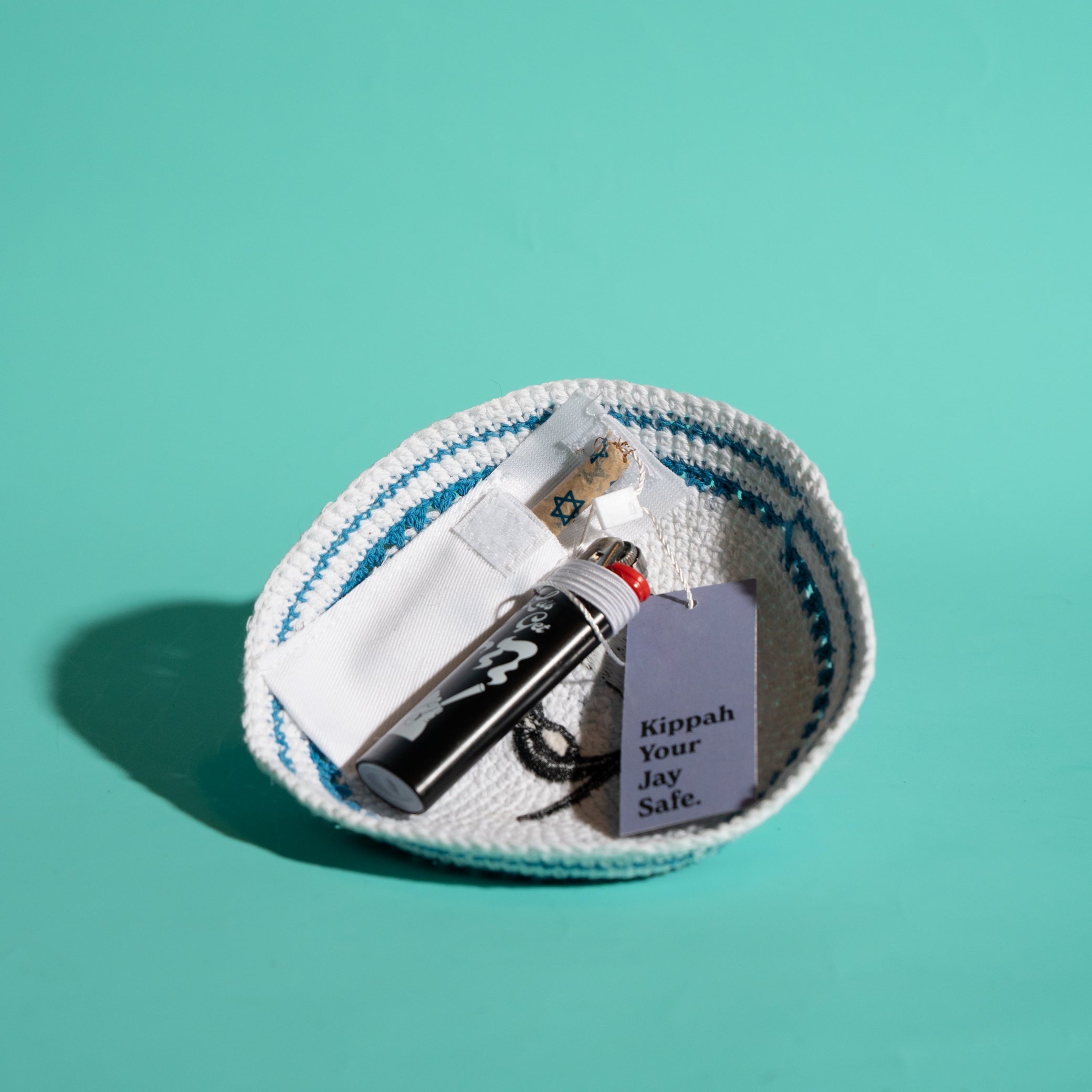 A small white knit kippah with blue stripes sits on a solid teal background, containing a black lighter, a joint holder, and a blue tag that reads 'Kippah Your Jay Safe'.