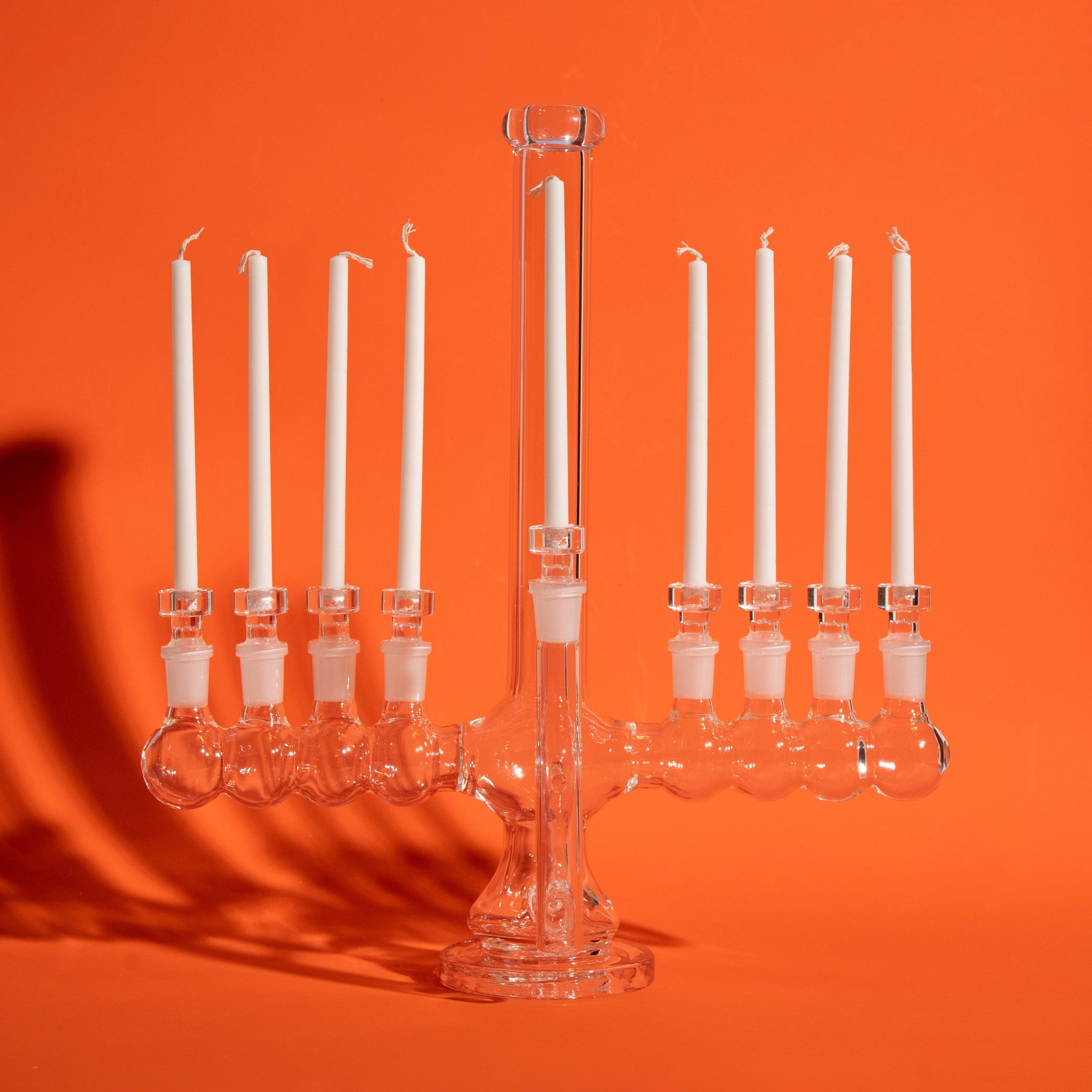 A clear glass menorah with nine white candles, set against a solid orange background with a soft shadow cast to the left.