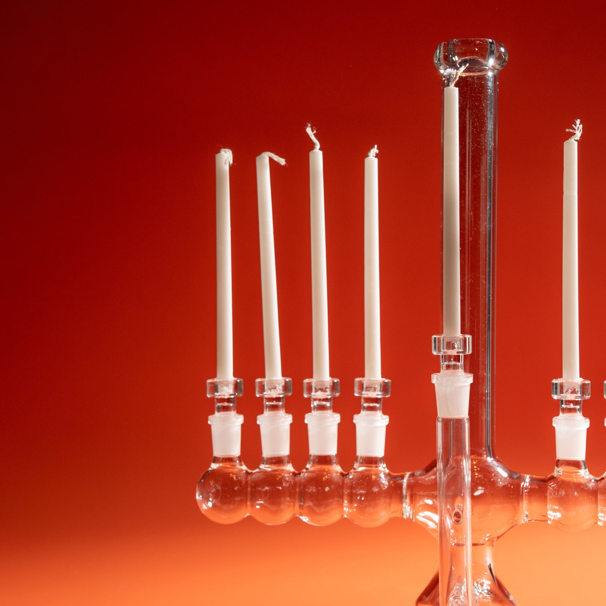 A clear glass menorah, photographed against a warm red-to-orange gradient studio background.