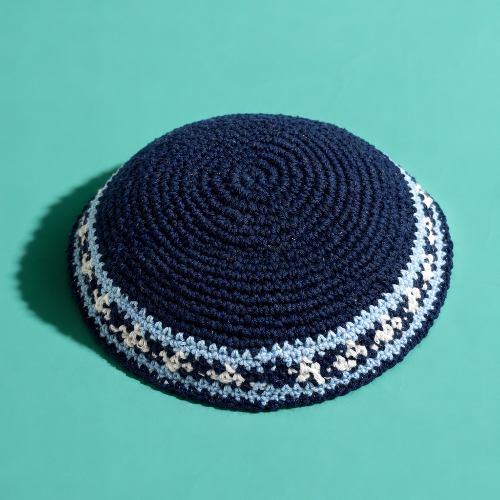 A dark navy blue crocheted kippah with light blue and white patterned trim, photographed on a solid teal background.