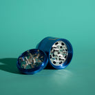 A blue metal herb grinder with a silver grinding mechanism, shown disassembled with the lid placed in front of the main body on a solid teal background.