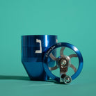 A blue metal herb grinder with a silver and blue hand crank, displayed on a solid teal background.