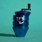 A blue metal herb grinder with a white "shin" on the side and a silver crank handle, set against a solid teal background.
