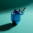 A metallic blue manual herb grinder with a silver handle, shown open on a teal shadowed background.