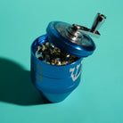 A blue metal herb grinder with a silver handle and lid, shown open and filled with dried herbs on a solid teal background.