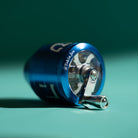 A close-up studio shot of a metallic blue grinder with a silver crank handle, resting on a teal surface against a dark teal background. A white "shin" is partially visible on the side.