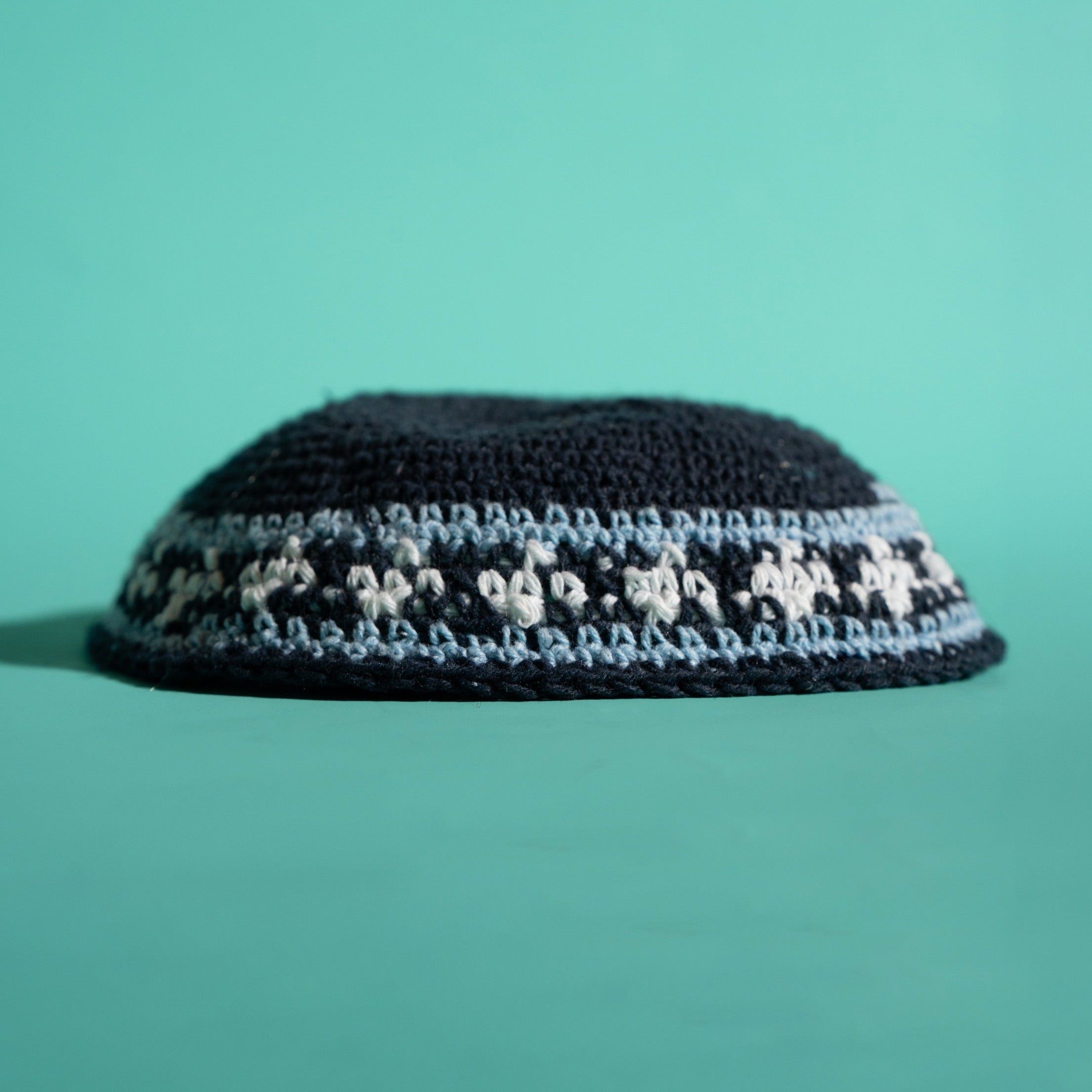 A black knitted kippah with a decorative band of white and light blue embroidered stars, set against a solid teal background.