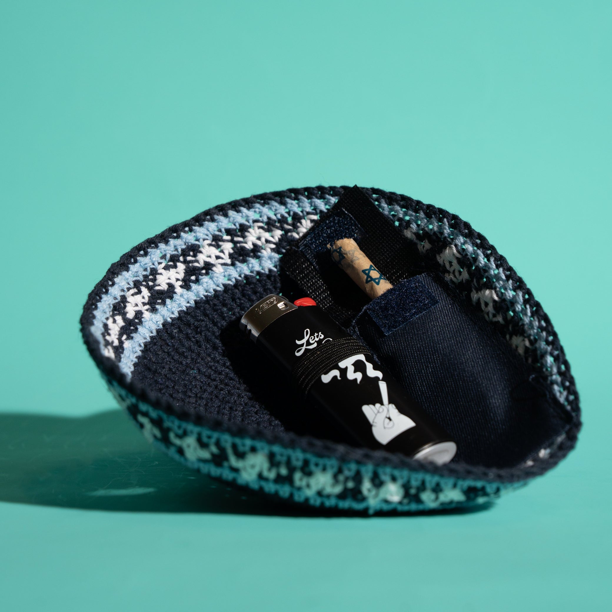 A small knit kippah in a dark navy blue color with a light blue and white knit pattern around the rim, shown on a solid teal background. Inside the pouch, a black lighter and a Tokin' Jay are visible.