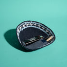 A knit kippah in navy blue with a white and light blue patterned trim, shown on a solid teal background. Inside the pouch, a black lighter and a small Tokin' Jay are placed.