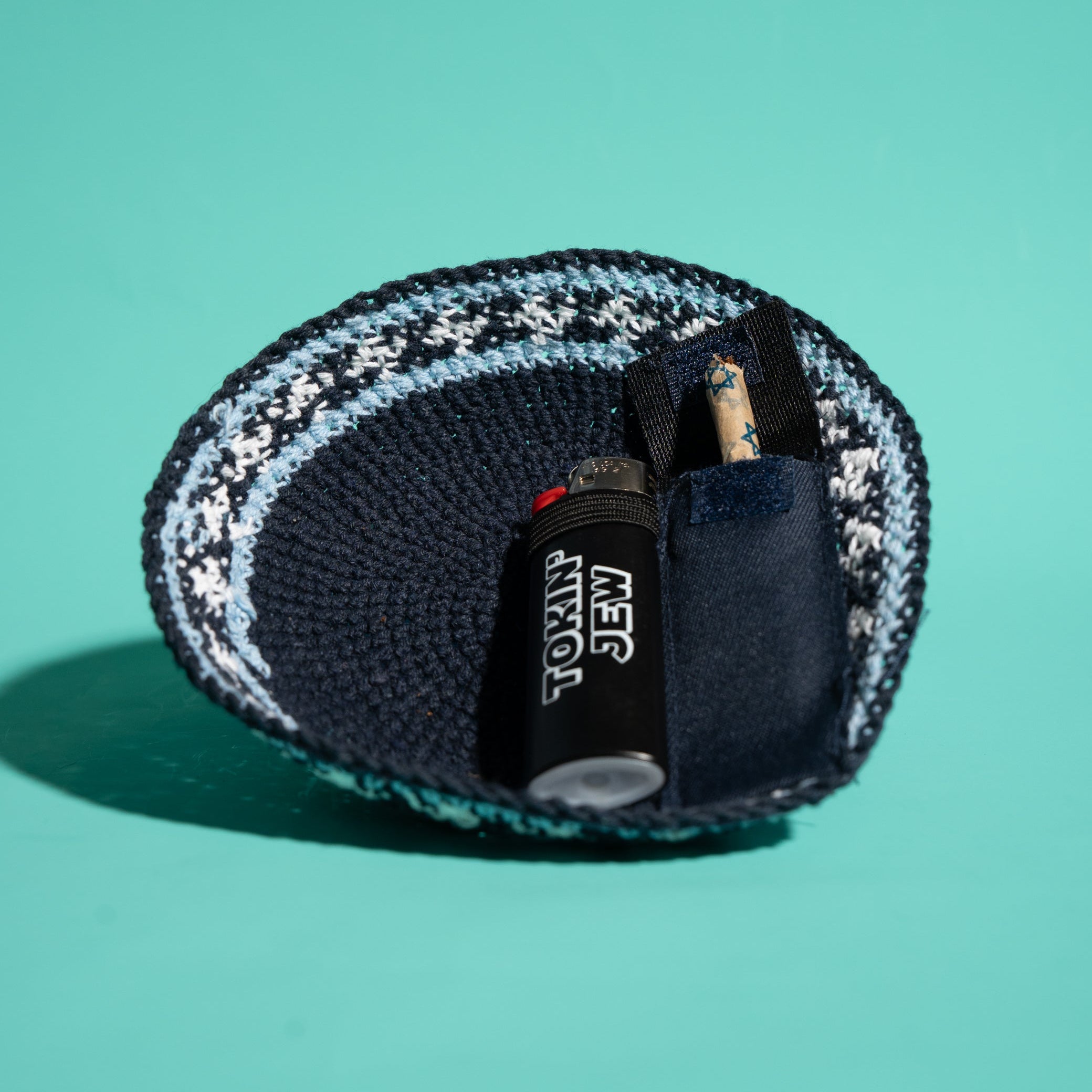 A knit kippah, dark navy, light blue, and white, shown on a solid teal background. Inside the pouch is a black lighter with white text reading 'TOKIN JEW' and a Tokin' Jay.