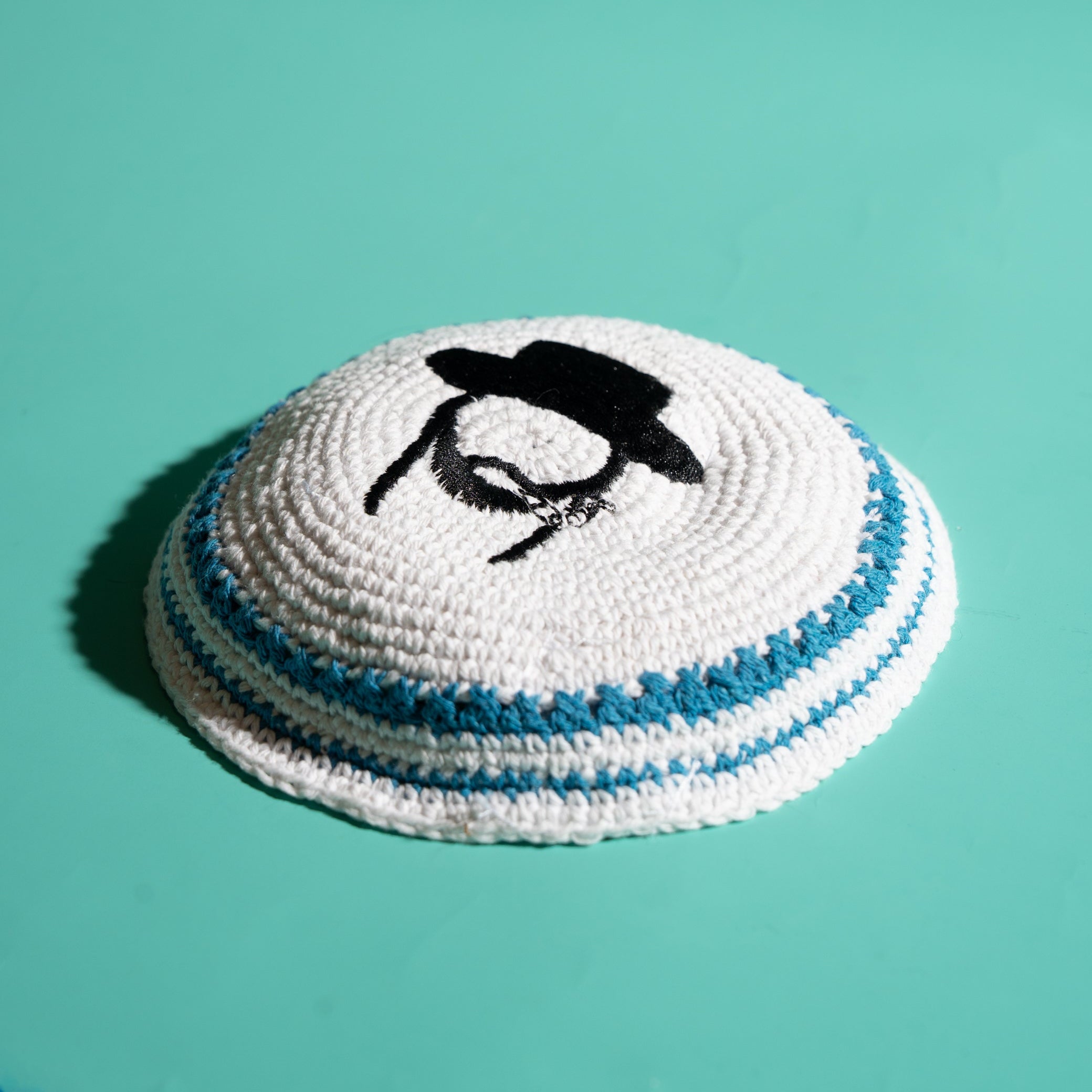 A white knit kippah with blue stripes and a black embroidered Rabbi design, shown on a solid teal background.