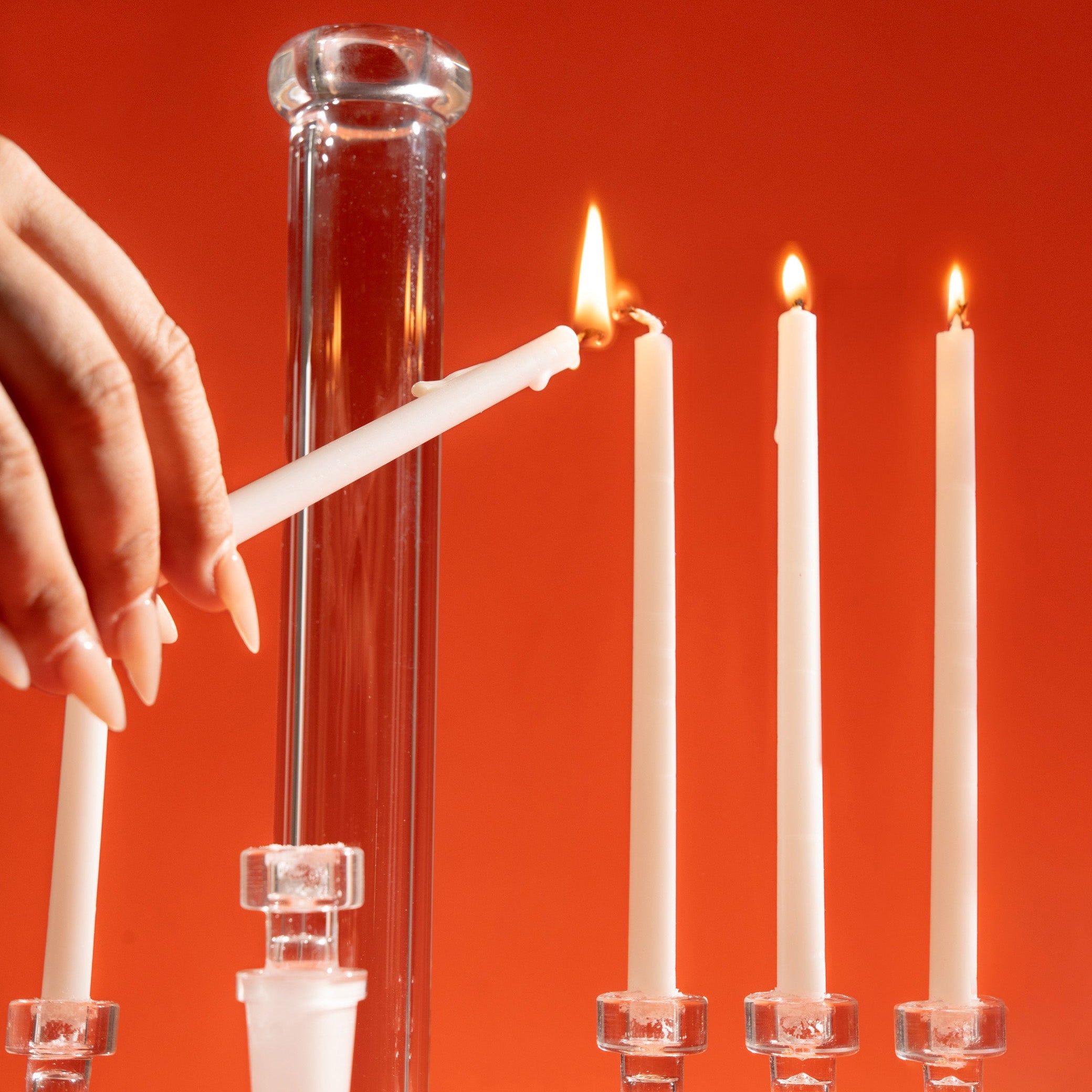 A hand lighting a white taper candle next to three more lit white candles standing in holders against a solid orange-red background. 
