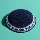 A dark navy blue crocheted kippah with light blue and white patterned trim, photographed on a solid teal background.