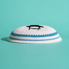 A white knit kippah with blue stripes and a black embroidered Rabbi on top, set against a solid teal background.