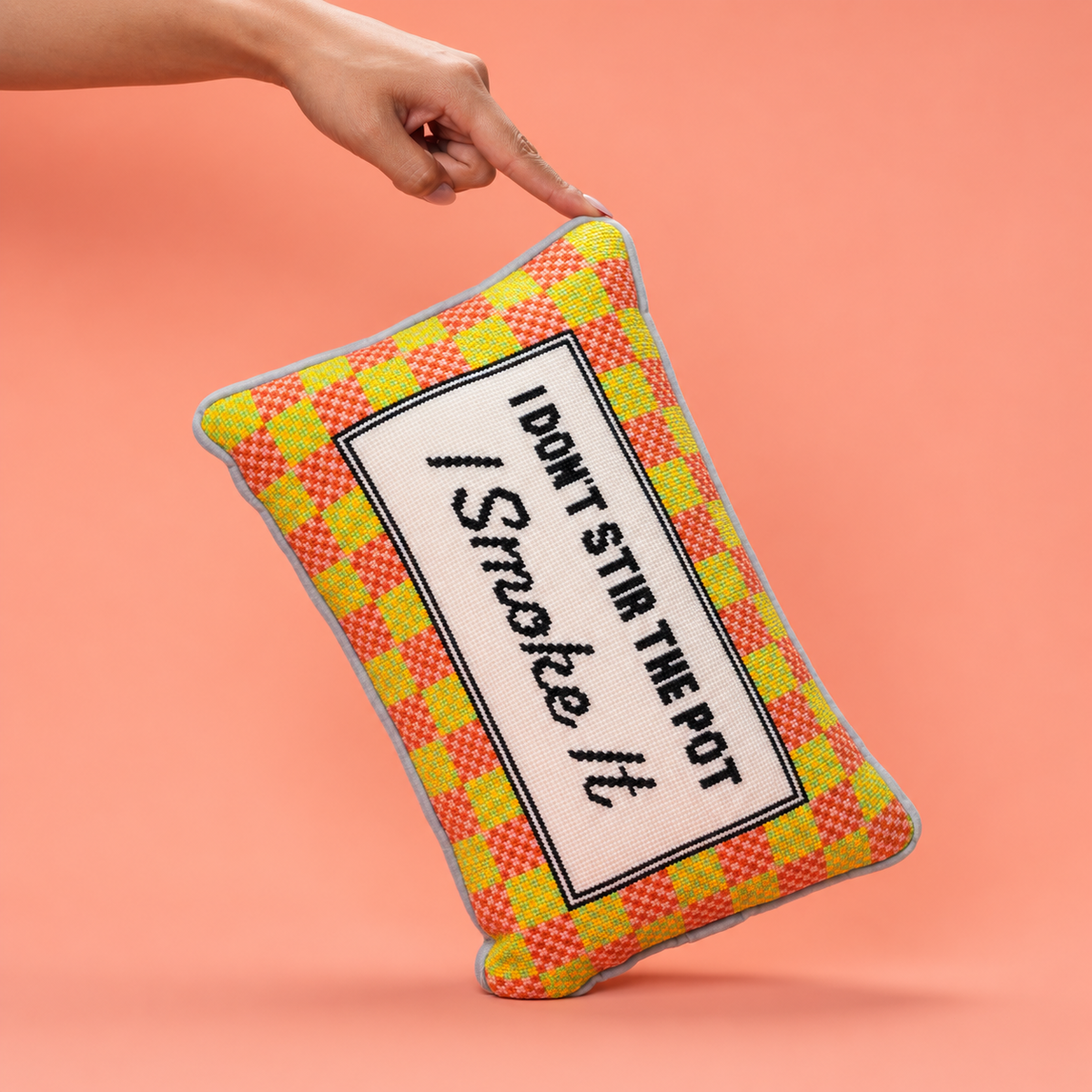 A rectangular decorative throw pillow with a yellow and red plaid pattern and a central white label reading 'I DON'T STIR THE POT / Smoke It', held by a person's hand against a solid peach background.