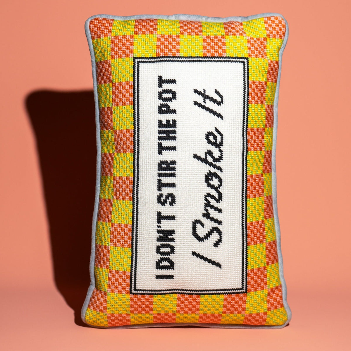 A rectangular decorative throw pillow with a yellow and orange checkered pattern border and a central white panel featuring the embroidered text 'I DON'T STIR THE POT / Smoke It'. The pillow is set against a solid peach-pink background with a soft shadow cast to the left.