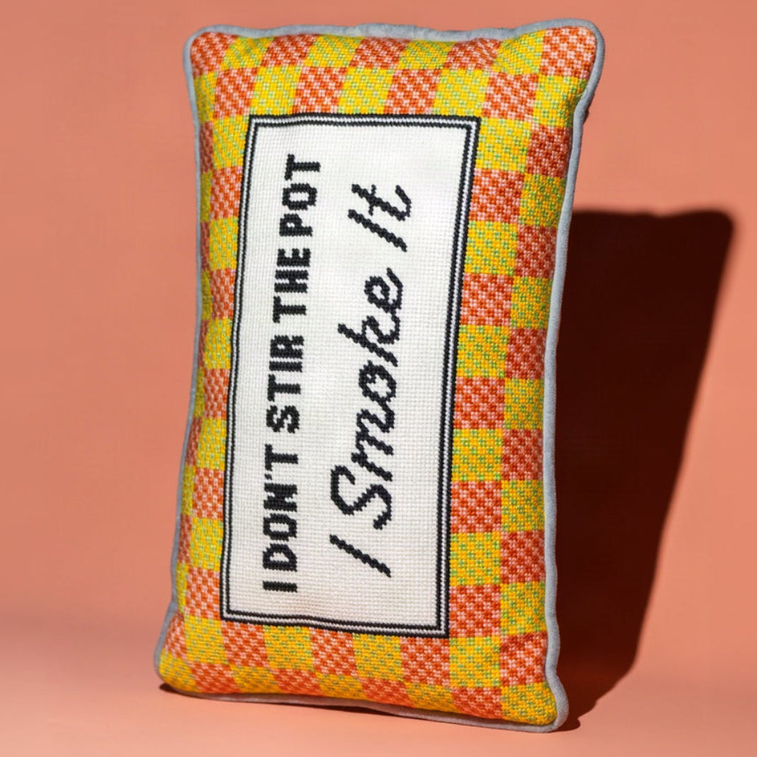A rectangular decorative pillow with a yellow and orange checkered pattern, featuring a white central panel with the embroidered text 'I DON'T STIR THE POT / Smoke It' in black. The pillow is photographed on a solid peach-pink background with a soft shadow cast to the right.