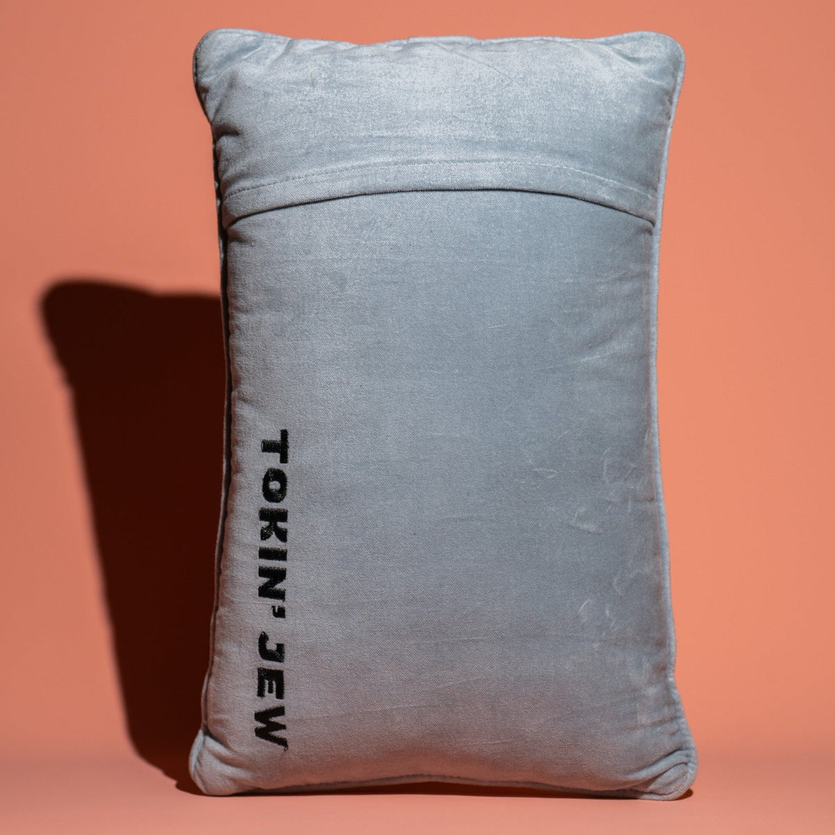 A light blue velvet rectangular pillow with the text 'TOKIN' JEW' printed vertically in black on the front, set against a solid peach background.