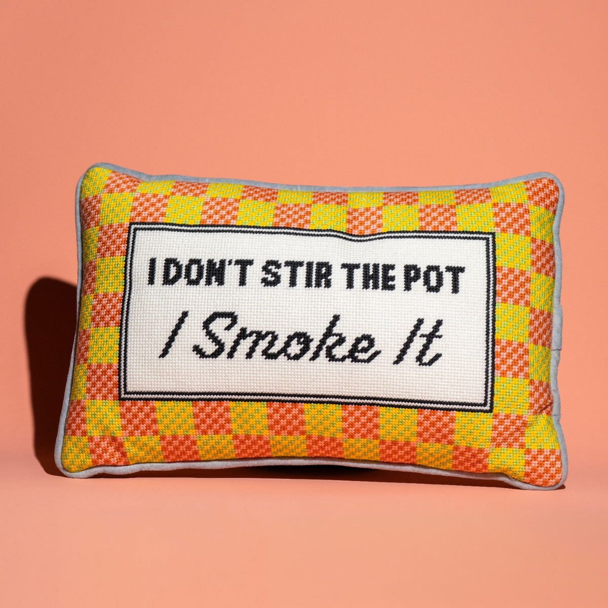 A rectangular decorative pillow with a yellow and orange checkered pattern, featuring a white central label with the embroidered text 'I DON'T STIR THE POT / I Smoke It'. The pillow is set against a solid peach-pink background with a soft shadow to the left.