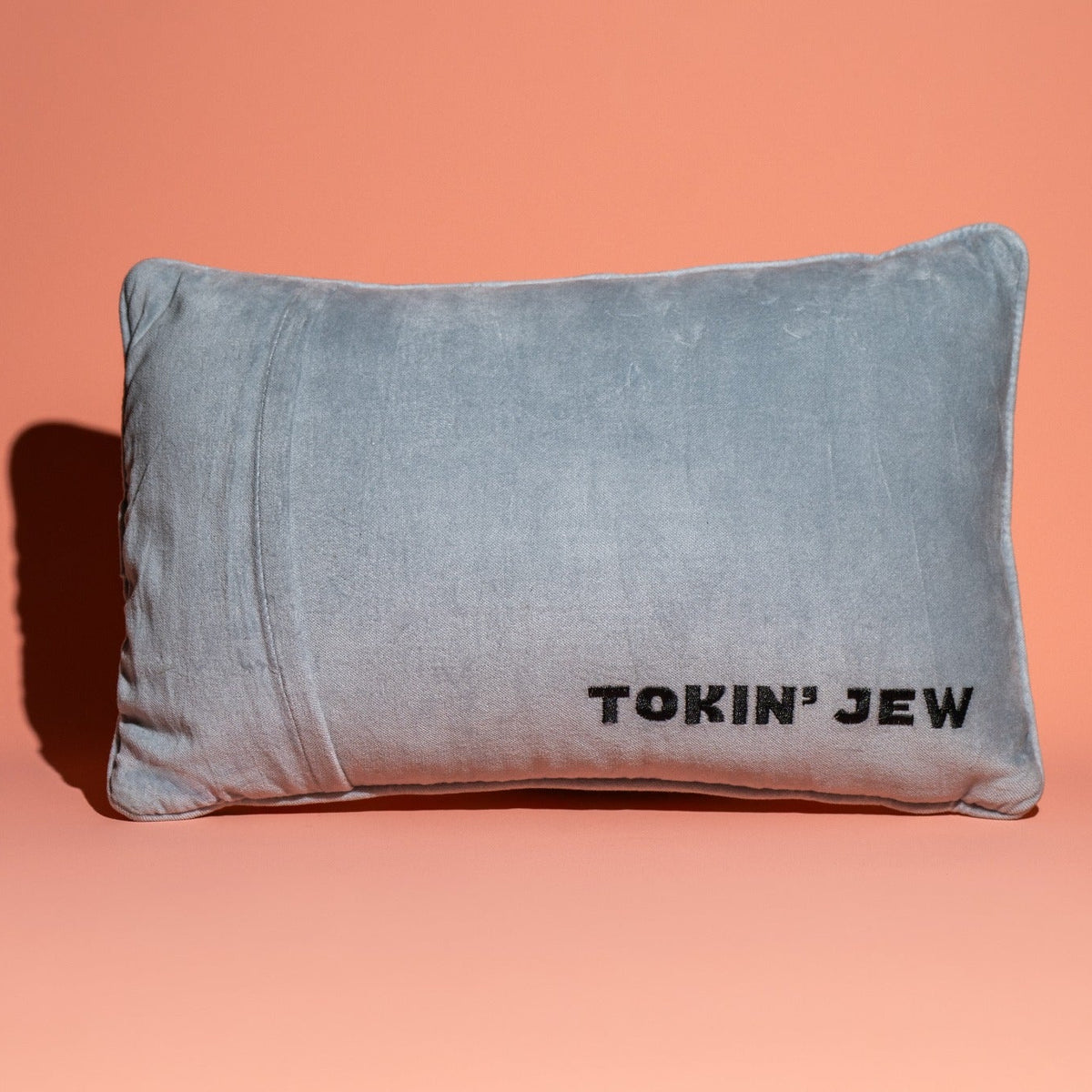 A rectangular light blue-gray velvet pillow with the text 'TOKIN' JEW' printed in black on the front, set against a solid peach-pink background.